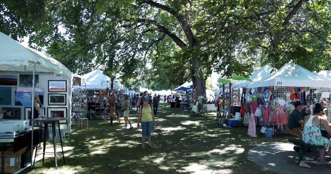 Visitors browsing rows of shaded arts and crafts booths beneath mature trees